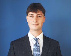 A smiling man with a cheque navy suit on with a light blue tie stood in front of a dark blue background