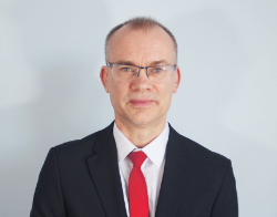 A smiling man with glasses and a black suit with a red tie stood in front of a light blue background