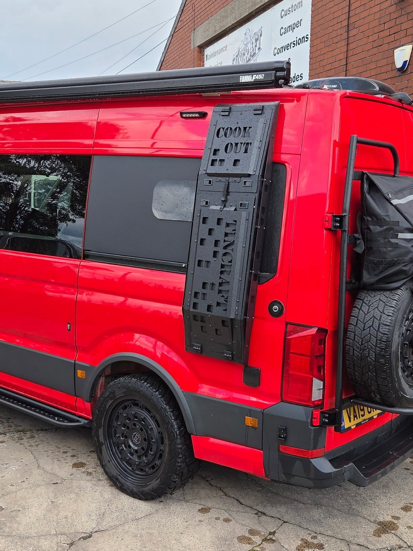 Rear three-quarter of a red VW Crafter overland camper with rear door storage panel and spare wheel