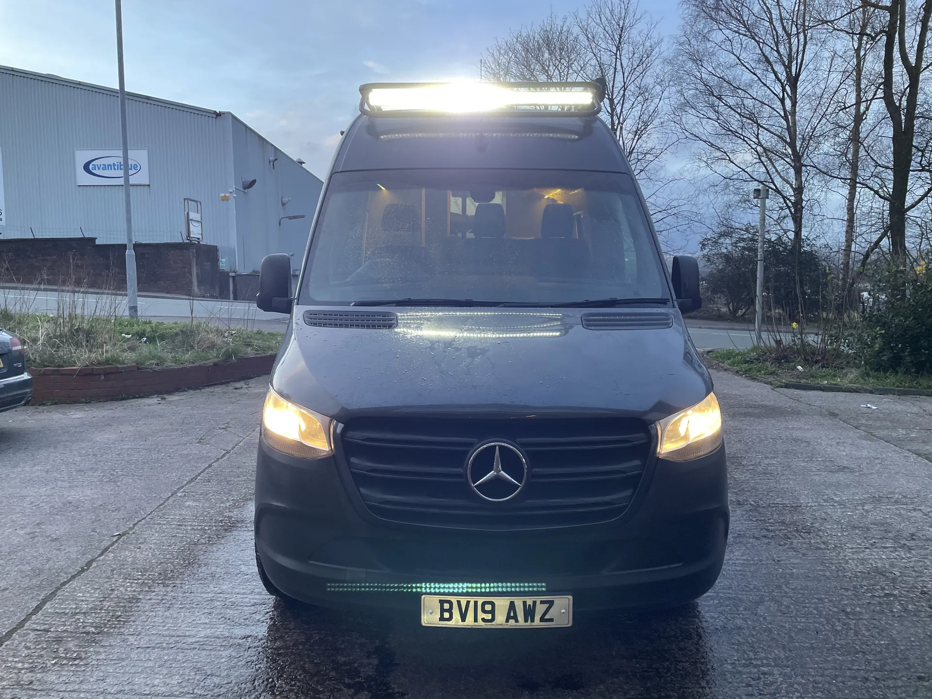 Front view of a dark grey Sprinter campervan at dusk with its headlights lit