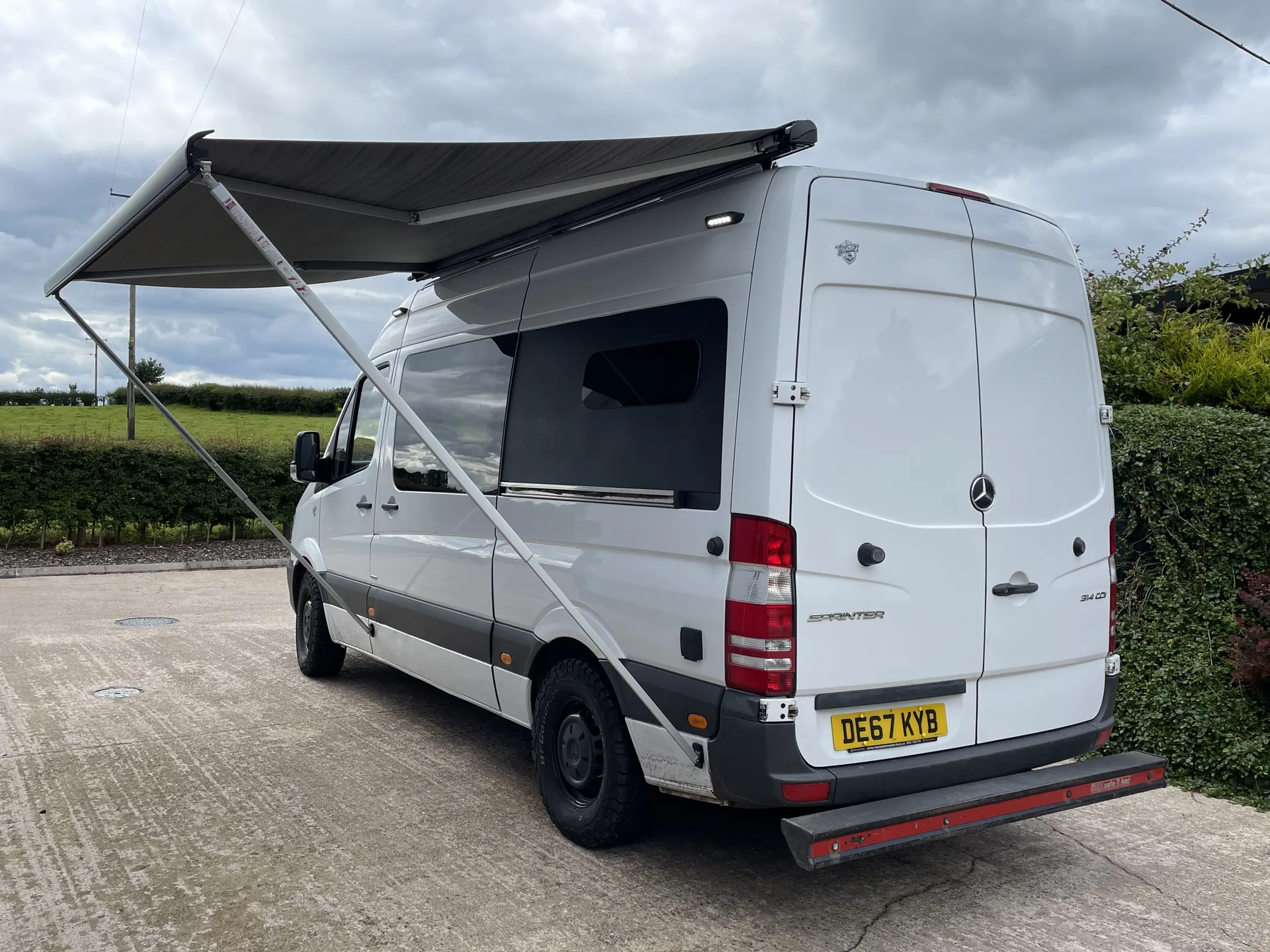 Rear three-quarter view of a Sprinter camper with its awning out in open countryside