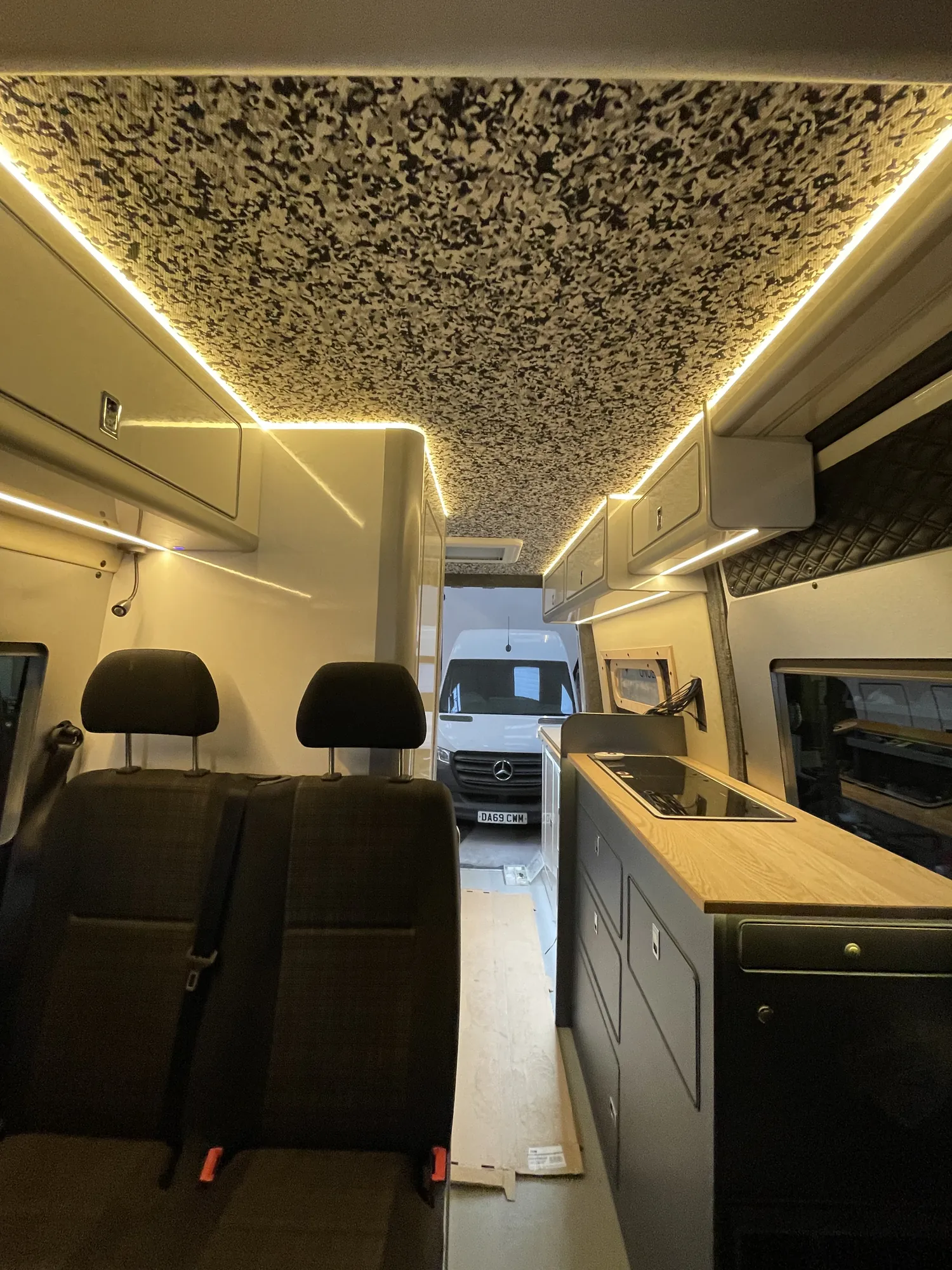 Sprinter camper interior looking forward past the seats to the galley, LED ceiling above