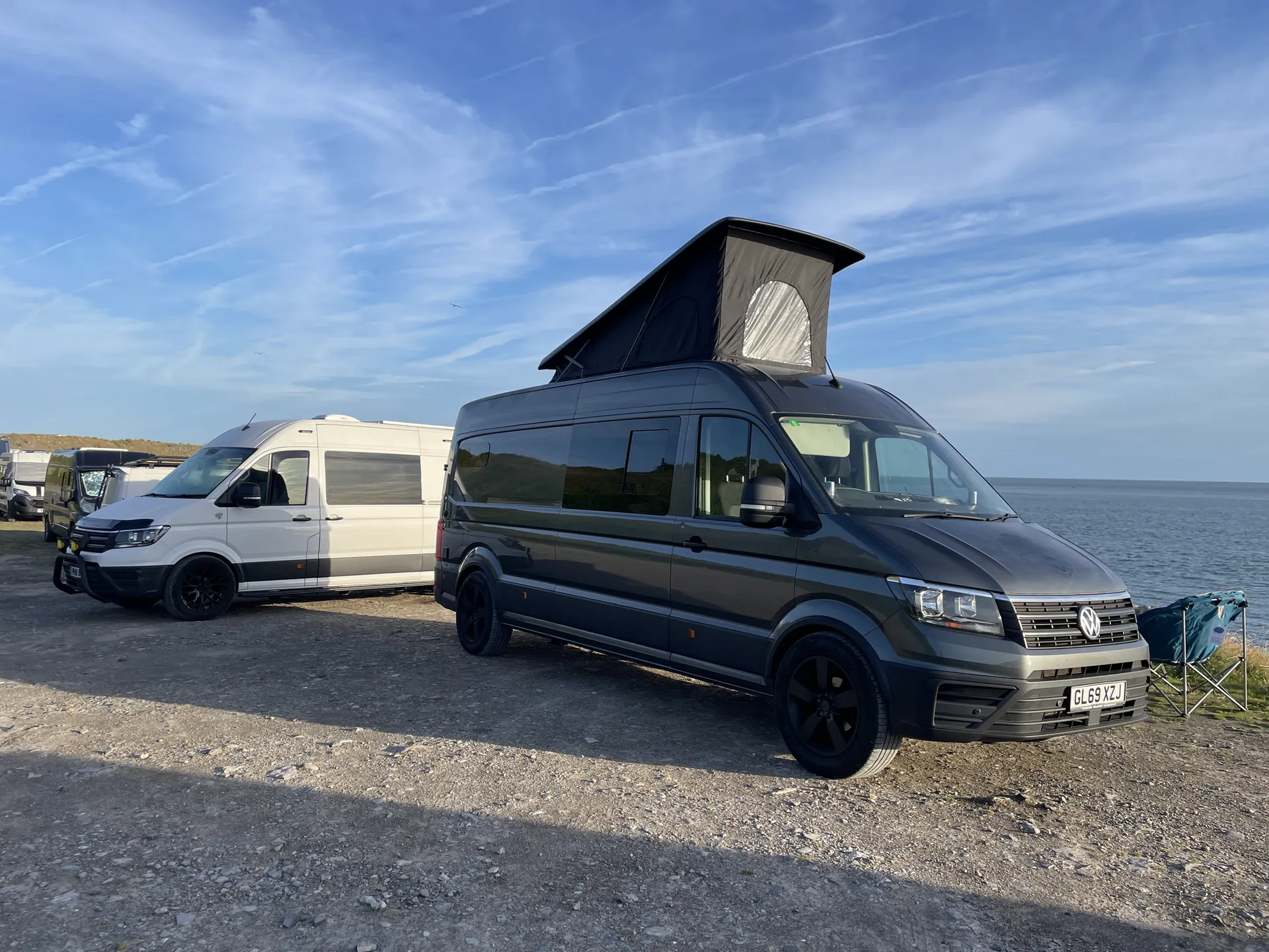 Grey VW Crafter camper with pop-top roof raised beside the coast under blue sky
