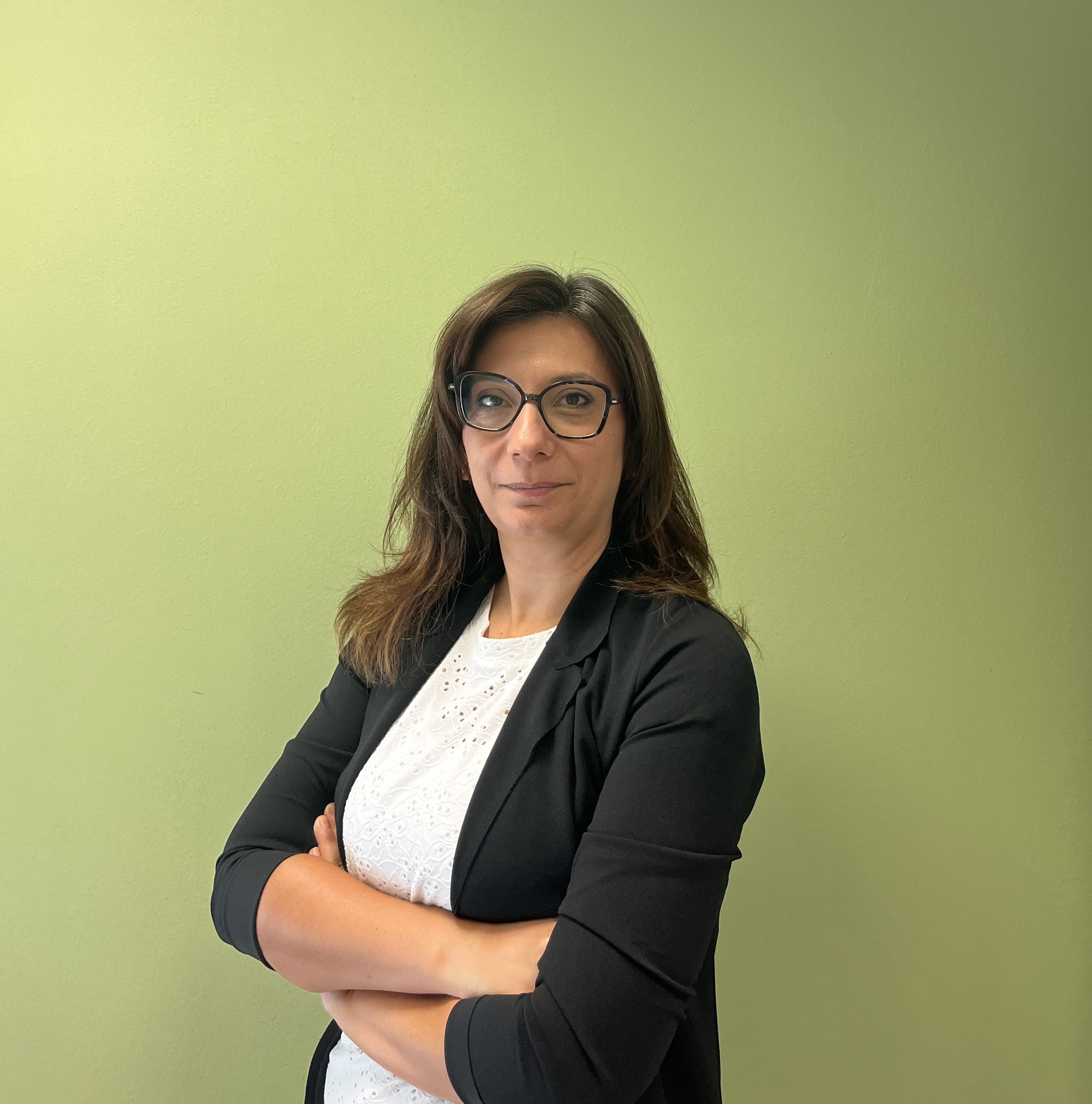 Donna in blazer nero e camicetta bianca, con le braccia incrociate davanti a un muro verde.