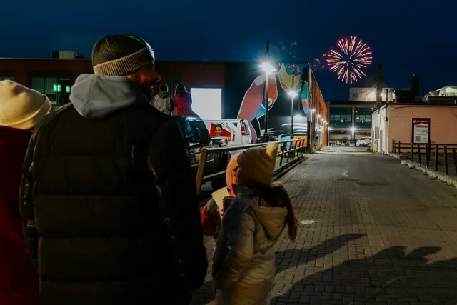 Fireworks burst over rooftops at night as bundled-up families watch from the street.