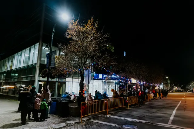 A crowd in winter coats lines a street strung with lights, waiting near a lit storefront at night.