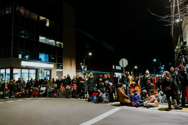 A large crowd gathers at a street corner at night, some seated on the curb with strollers.