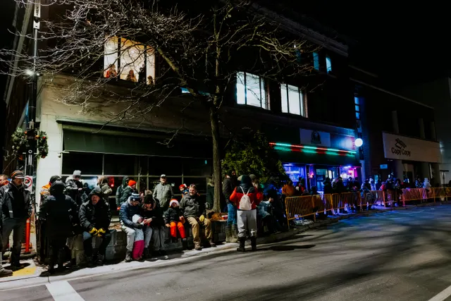 Spectators sit and stand along a row of storefronts strung with lights, waiting at night.