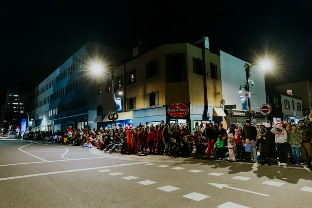 A crowd bundled in winter clothing waits along a storefront strip lit by streetlamps at night.