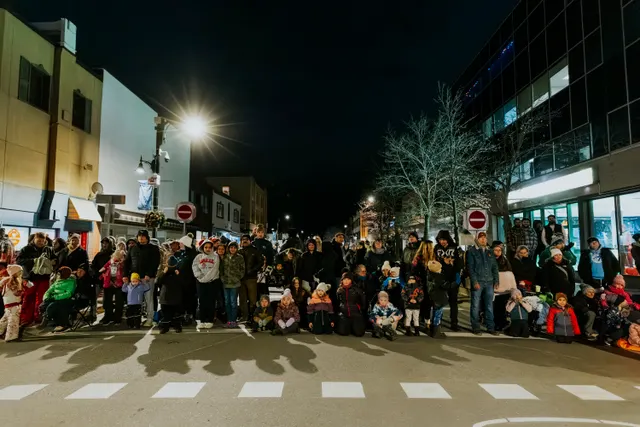 A crowd fills a crosswalk and both sidewalks of an empty, streetlamp-lit street at night.