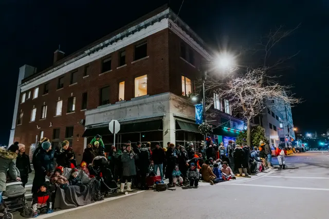 Spectators gather on a street corner beneath a brick building’s lit window at night.