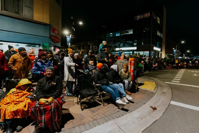 A crowd waits at a street corner, adults and children bundled up, some seated on camp chairs.