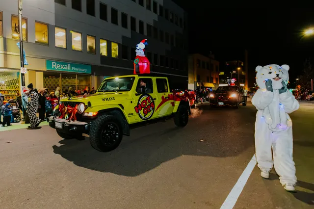 A yellow Jeep decorated like a hot dog leads the parade past a Rexall Pharmacy storefront.