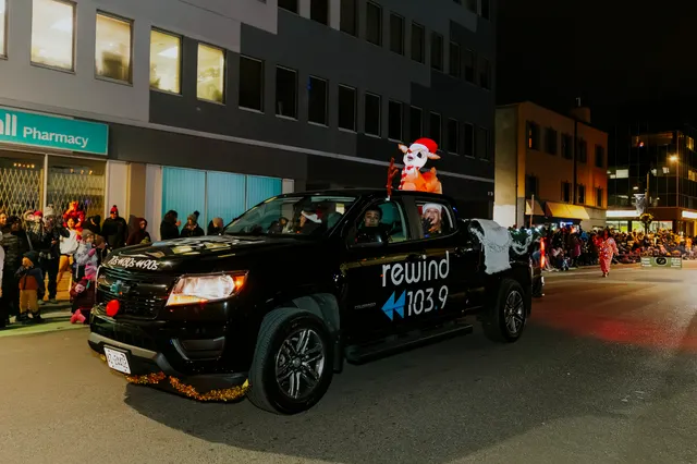 A black pickup truck branded Rewind 103.9 drives the parade route past a crowd on the sidewalk.