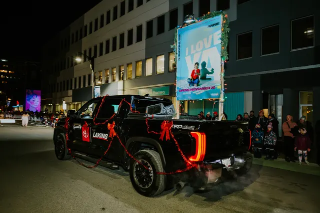 A black pickup truck decorated with red ribbon carries a lit sign advertising a November event.