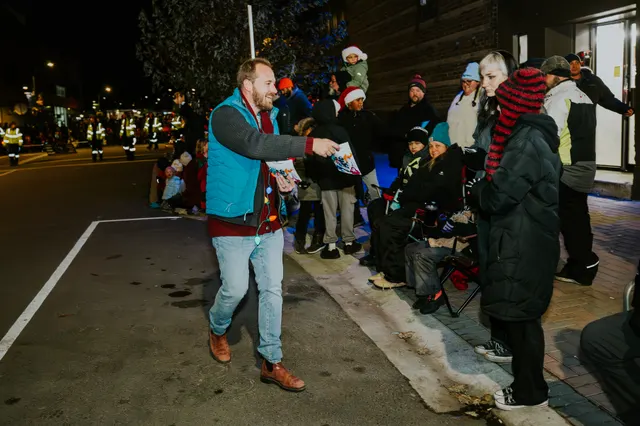 A man in a blue vest hands out treats to spectators lining a sidewalk at night.