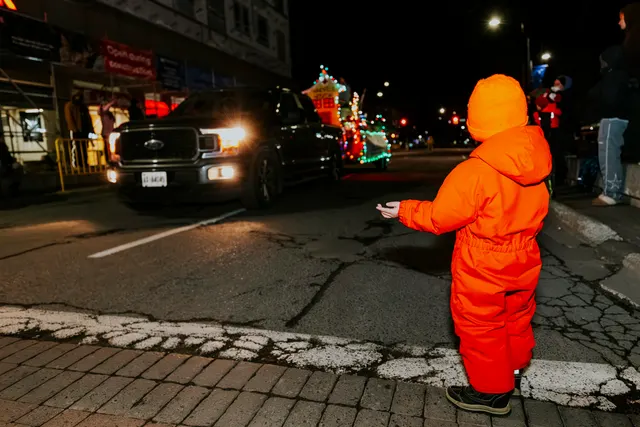 A child in an orange snowsuit reaches toward a pickup truck leading a lit float down the street.