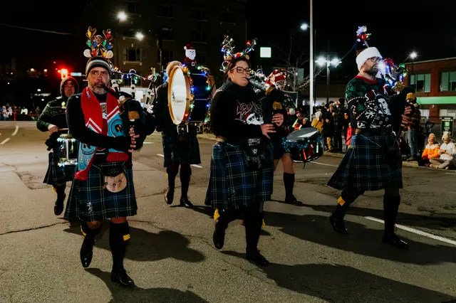 Kilted pipers and drummers wearing lit antler headbands march in the parade at night.