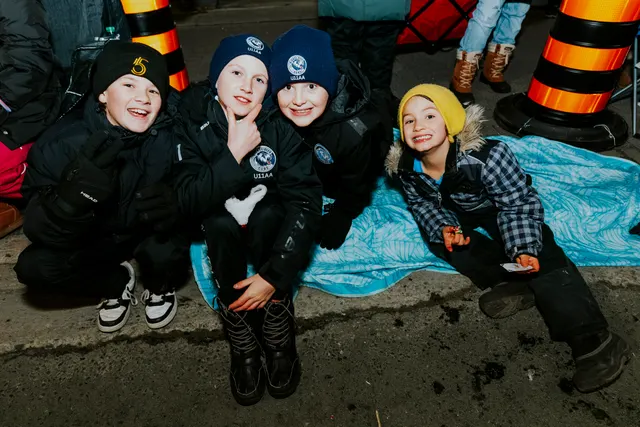 Four children sit together on a blanket at the curbside, smiling at the parade at night.