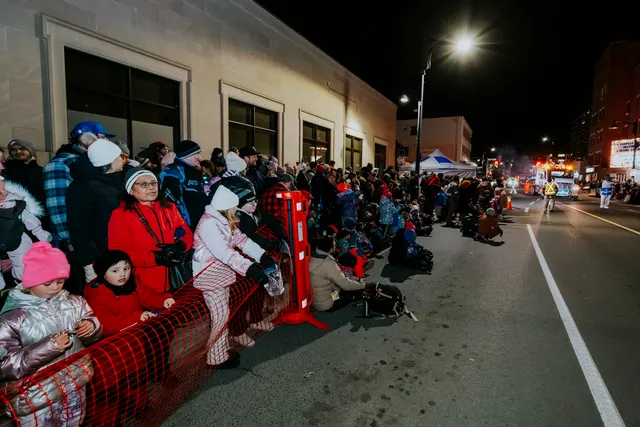Spectators line a red barrier as a lit parade truck approaches in the distance.