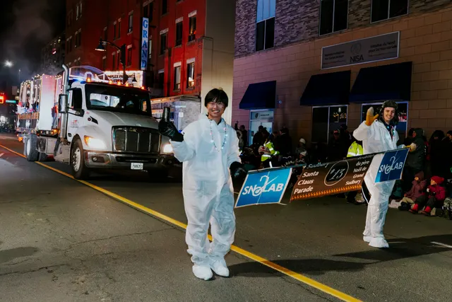 Two walkers in white coveralls give thumbs up, carrying SNOLAB parade banners past a truck.