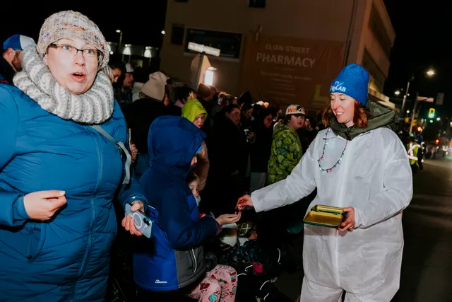 A SNOLAB-costumed marcher hands a treat to a child as spectators watch outside a pharmacy.