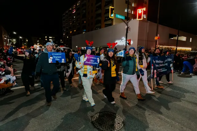 Marchers in antler headbands carry bilingual holiday greeting signs through an intersection at night.
