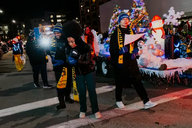 People in striped scarves stand beside a float decorated with a lit Christmas tree and a snowman.