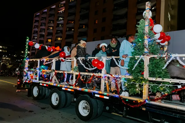 A flatbed float decorated with garland, balloons and Christmas trees carries riders past an apartment building.