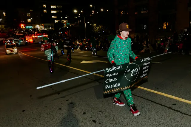 A performer in an elf costume rides a unicycle beside a Sudbury Santa Claus Parade entry sign.