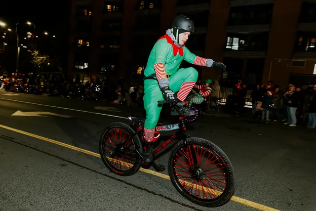 A rider in an elf costume balances a wheelie on a bike with red-lit wheels.