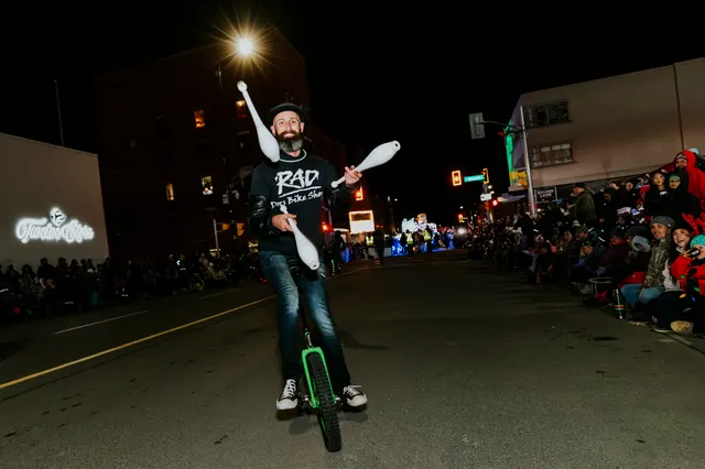 A man juggles clubs while riding a unicycle down the parade route at night.