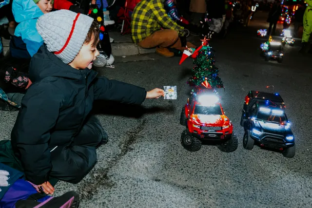 A child reaches for a treat beside two toy trucks decorated with a lit miniature Christmas tree.