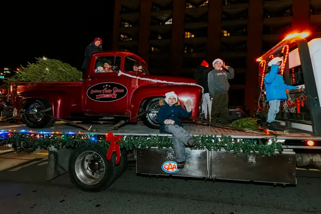 Children ride a flatbed trailer carrying a vintage red pickup truck loaded with a Christmas tree.