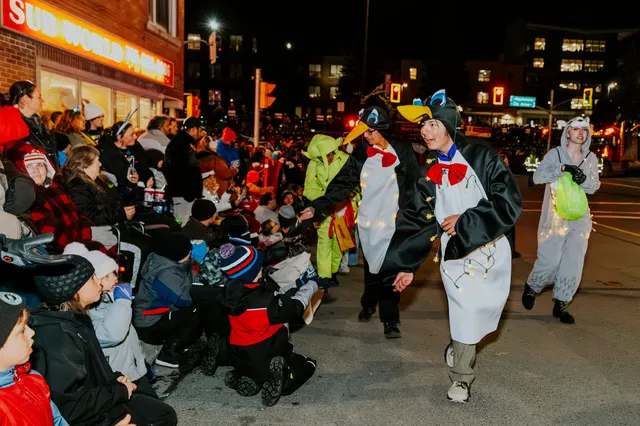 Performers in penguin costumes dance and greet a crowd seated along the sidewalk at night.