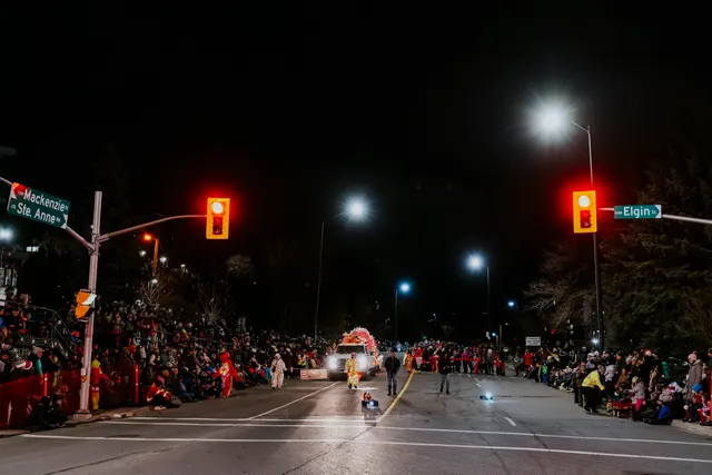 A lit float approaches an intersection marked by red traffic signals as crowds line both sides.