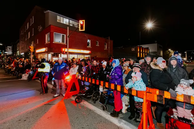 A crowd waits behind an orange barrier as a marshal with a flashing light stands watch.