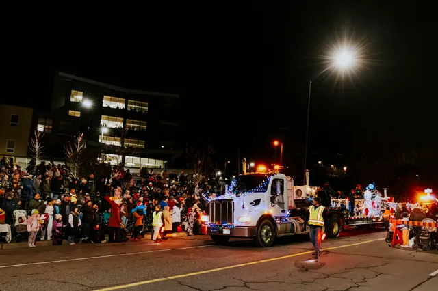 A white semi-truck tows a lit float past bleachers full of spectators at night.