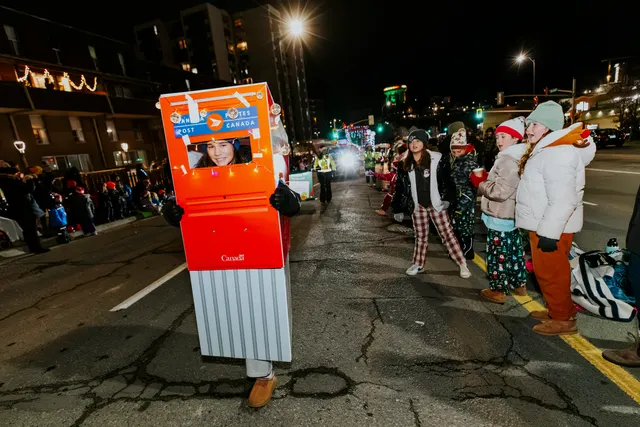 A person wears a large Canada Post mailbox costume, walking past spectators in pajamas.