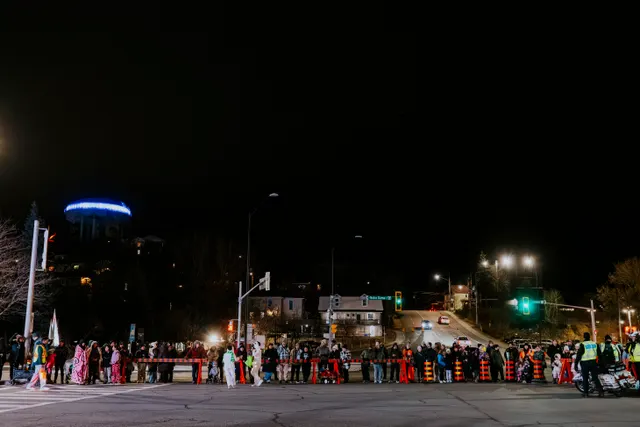 Spectators line both sides of an intersection at night beneath a glowing blue dome in the distance.