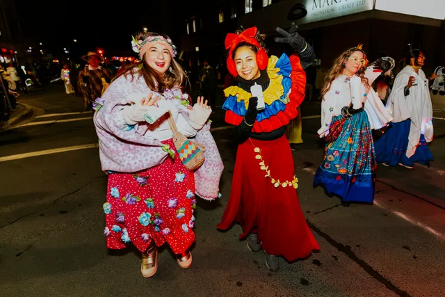 Two women in colorful ruffled dresses walk the parade route surrounded by other costumed dancers.