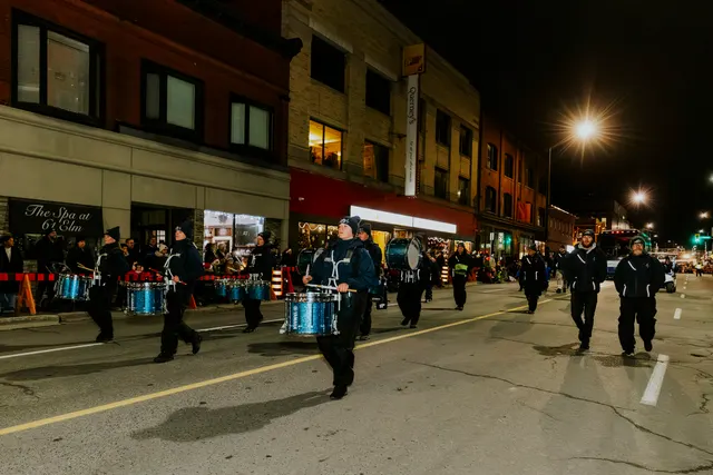 A marching band plays blue drums down the street past storefronts at night.