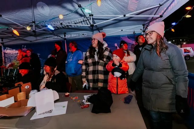 Volunteers and families gather under a light-strung tent at a parade night booth.