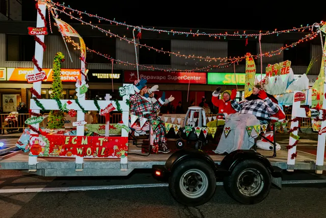 Costumed performers wave from a whimsical, brightly lit float decorated with holiday bunting.