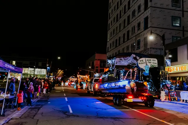 A narwhal-themed float with a sign reading Bye Buddy Hope You Find Your Dad passes a lit crowd.