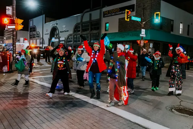 A group in festive light-up outfits cheers with arms raised at a street intersection.
