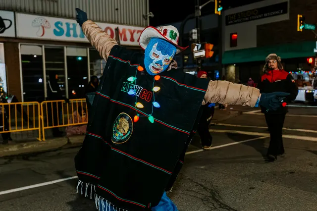 A performer in a blue lucha libre mask and a poncho reading Mexico dances down the street.