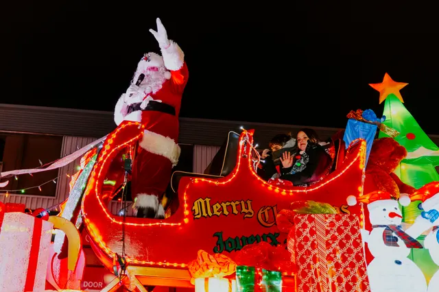 Santa waves from a red sleigh float outlined in lights, with 'Merry Christmas' spelled along its side.