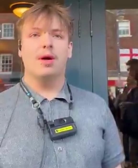 The manager, wearing a body-worn camera device, speaks to customers at the pub entrance as an England flag is visible among the crowd gathered outside behind him.