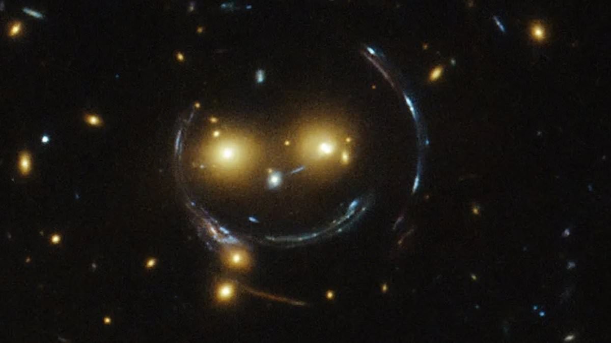 A NASA image showing a giant "cosmic smile" shape formed by warped spacetime, a phenomenon caused by gravitational lensing bending light from distant galaxies.